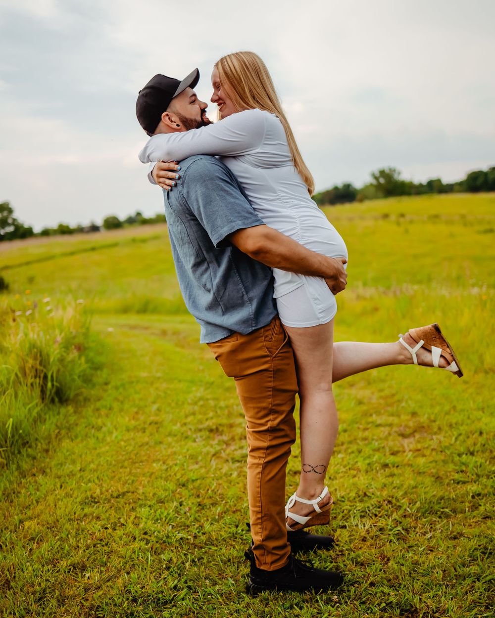 A couple embraces in a romantic pose on a grassy field during a sunny day with trees in the background.