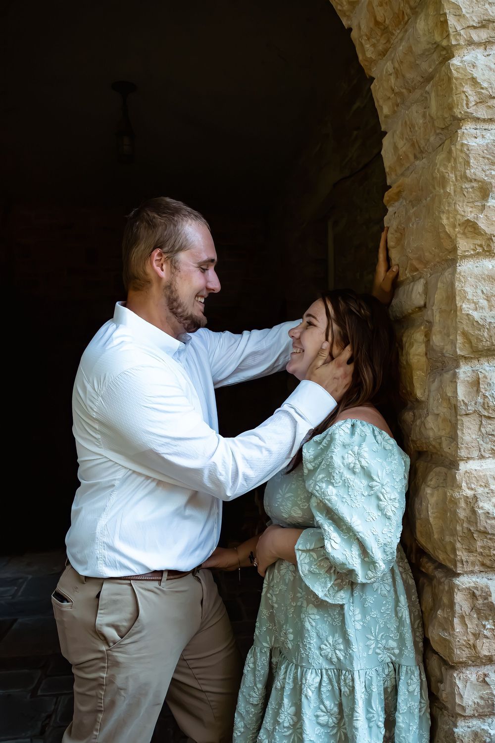 A couple shares a tender moment in a stone archway, with the person in white touching their partner wearing a mint dress.