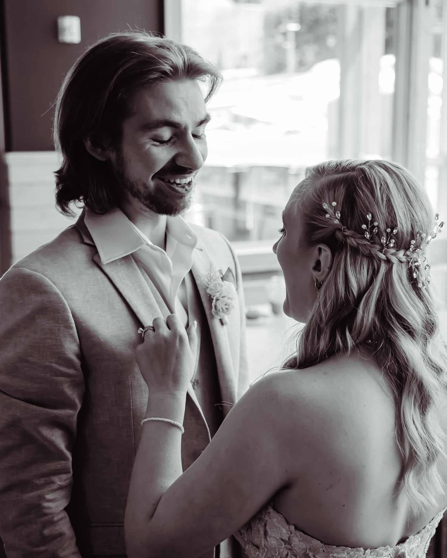 Newlyweds share an intimate black and white moment as couple embraces near window in formal wedding attire.
