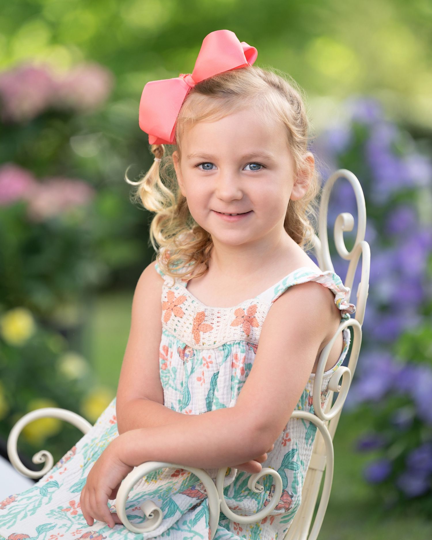 A child wearing a pink dress and necklace sits on a white decorative chair in a sunny garden setting.