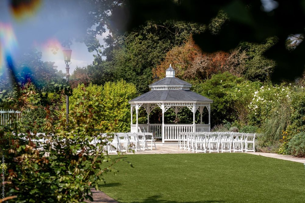 White wooden gazebo set up for outdoor wedding ceremony with rows of chairs on manicured lawn surrounded by garden.