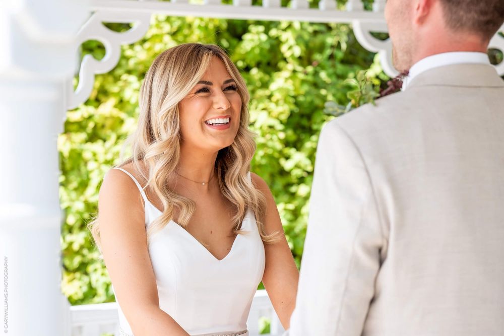 Bride in white dress laughs joyfully during outdoor wedding ceremony under leafy green canopy.