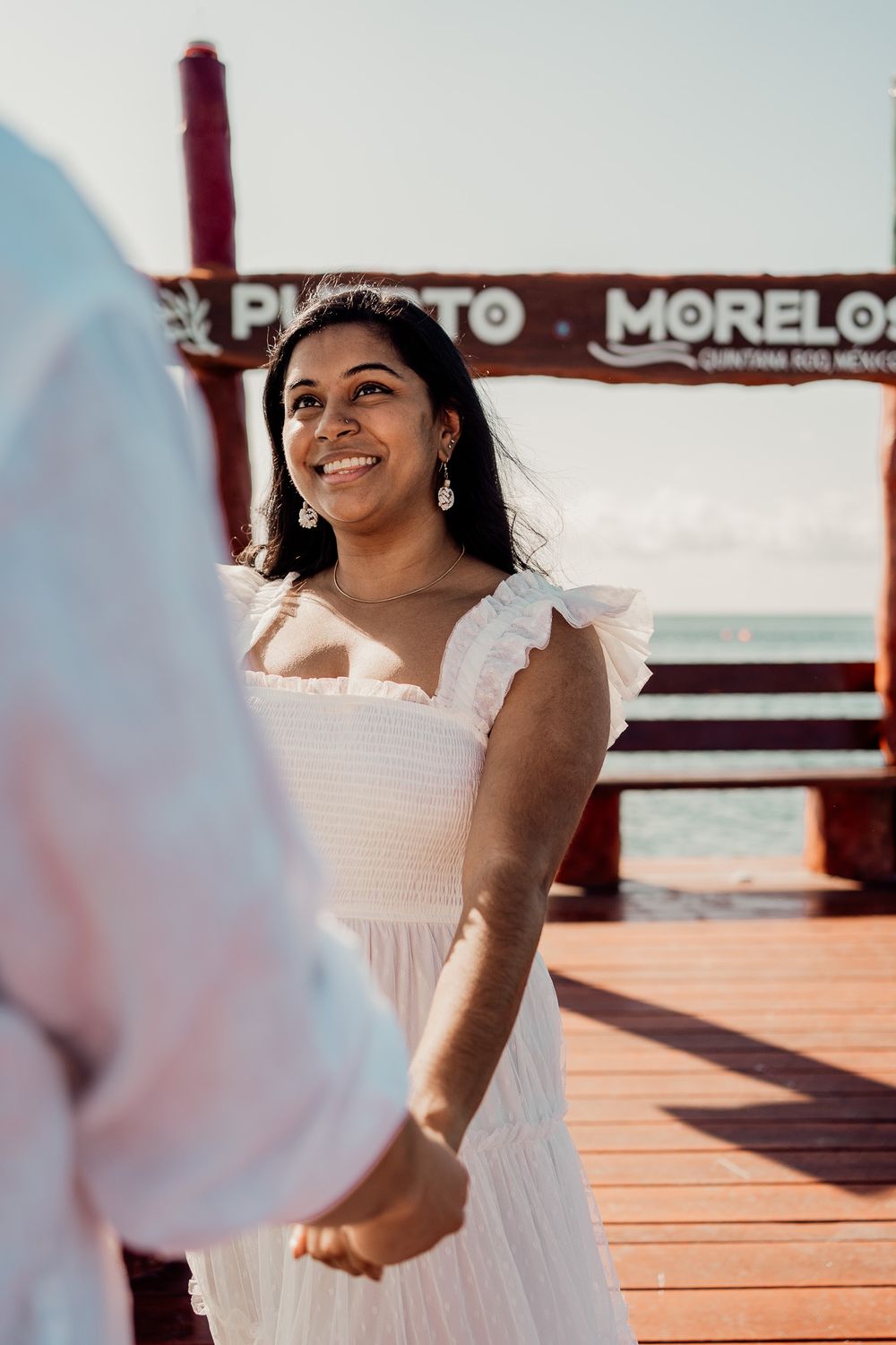 Woman in white dress smiling brightly on wooden pier with Puerto Morelos sign photoshoot in Puerto Morelos.