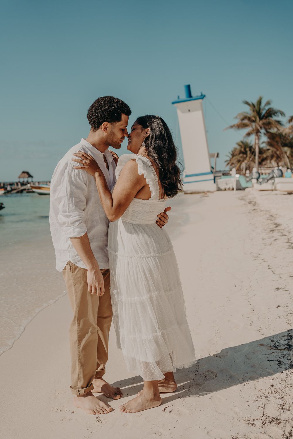 Couple in white outfits sharing intimate moment on sunny beach with palm trees photoshoot in Puerto Morelos.
