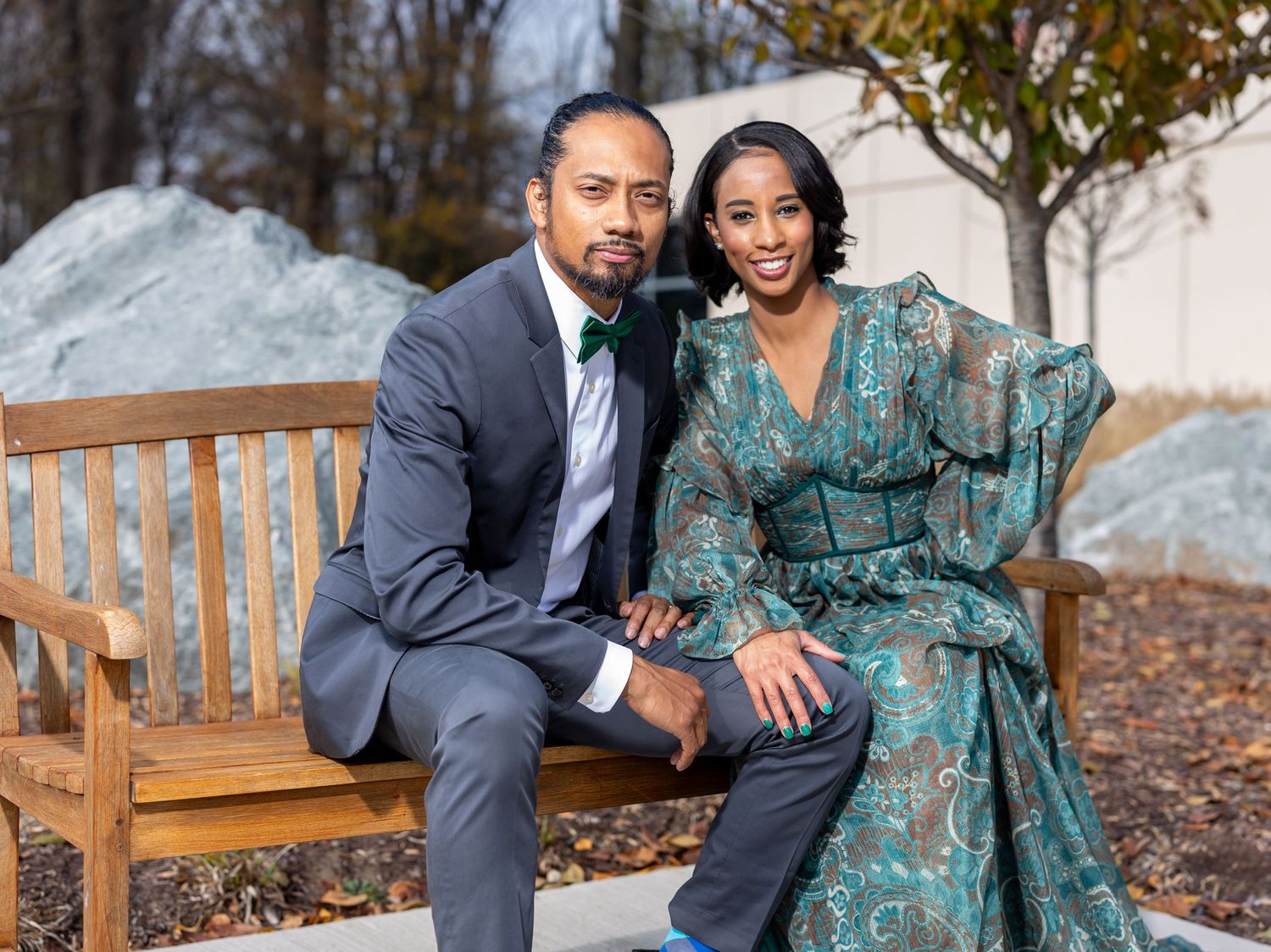 A couple sits together on a wooden bench outdoors in formal attire.