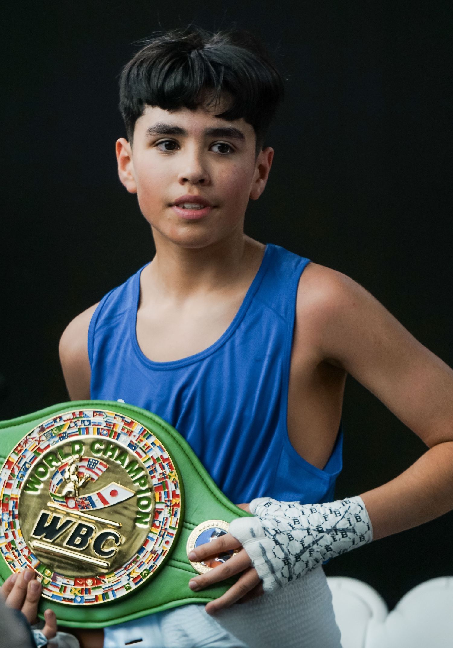 Young boxer in blue tank top poses with green and gold WBC championship belt against dark background.