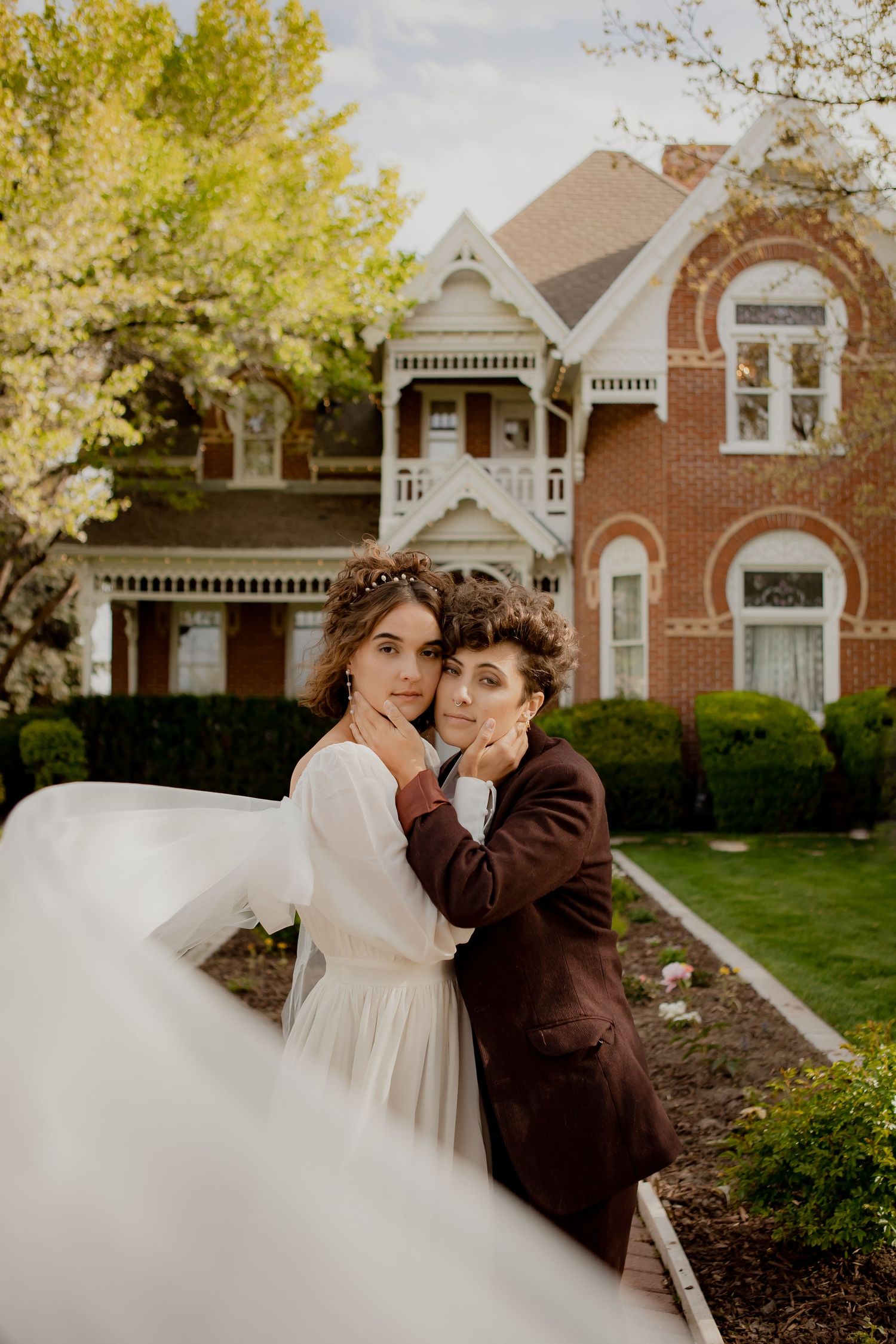 A romantic wedding portrait in front of a Victorian brick mansion with blooming trees.