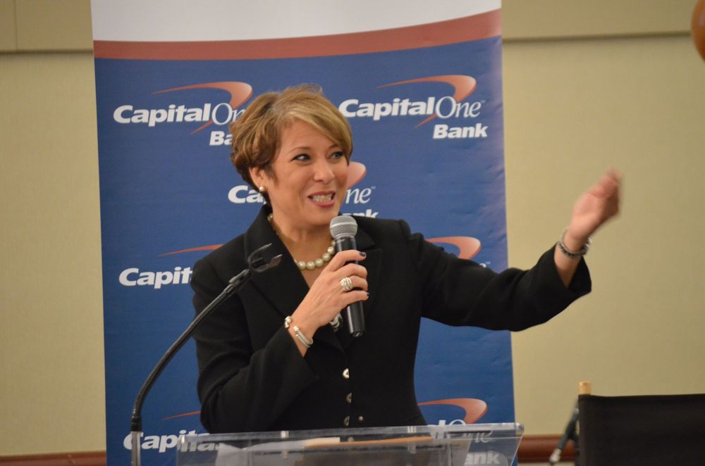 Woman speaking at Capital One Bank event, gesturing expressively at podium with microphone.