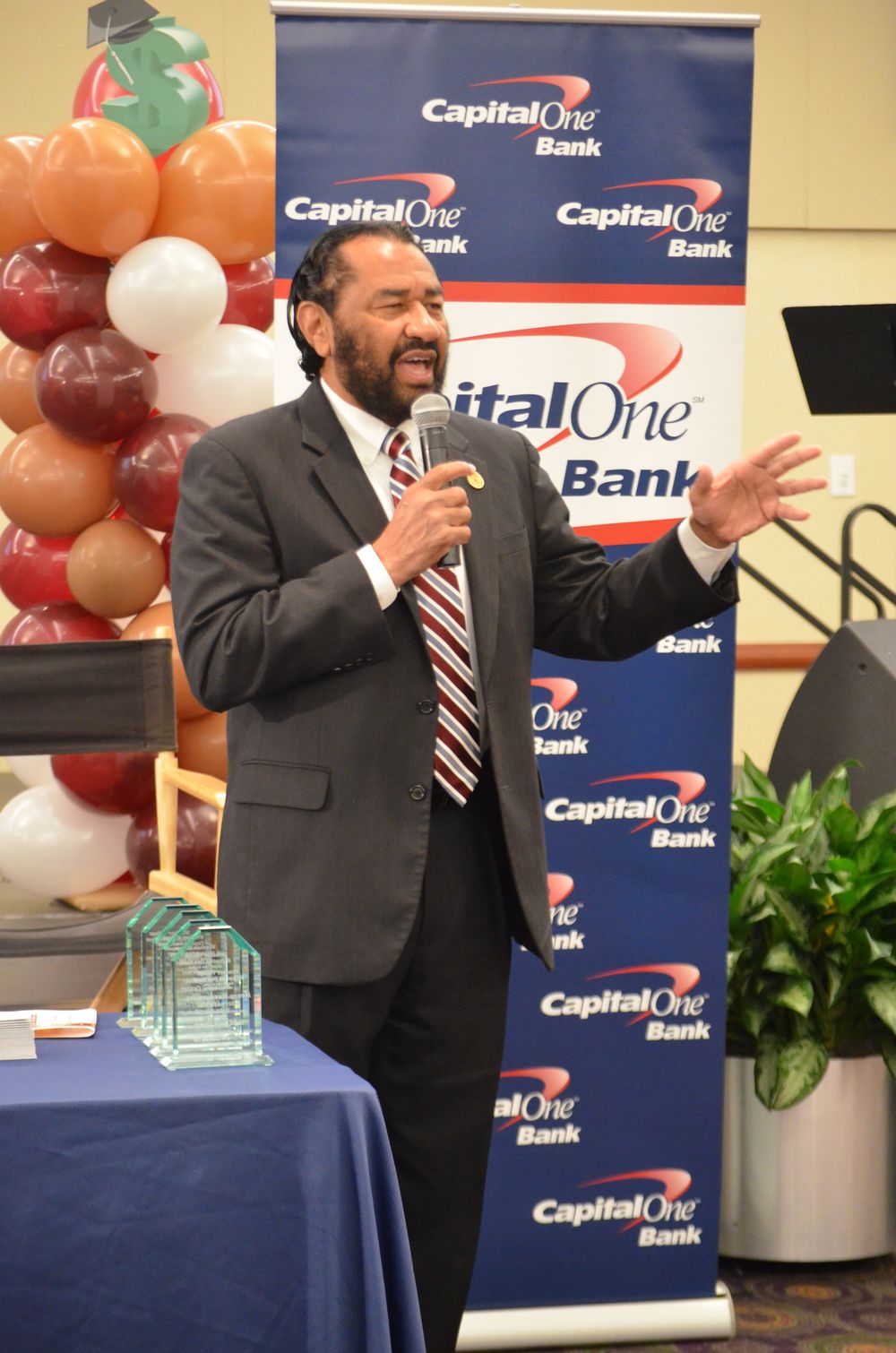 Man speaking at Capital One Bank event, holding microphone, gesturing with hand, awards on table, balloons in background.