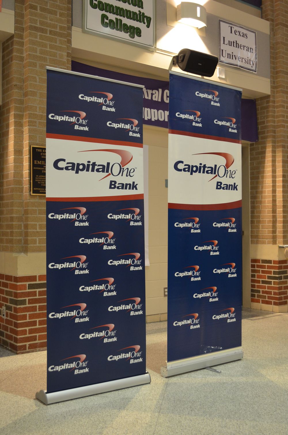 Two Capital One Bank promotional banners displayed in a brick building lobby near community college and Texas Lutheran University signs.