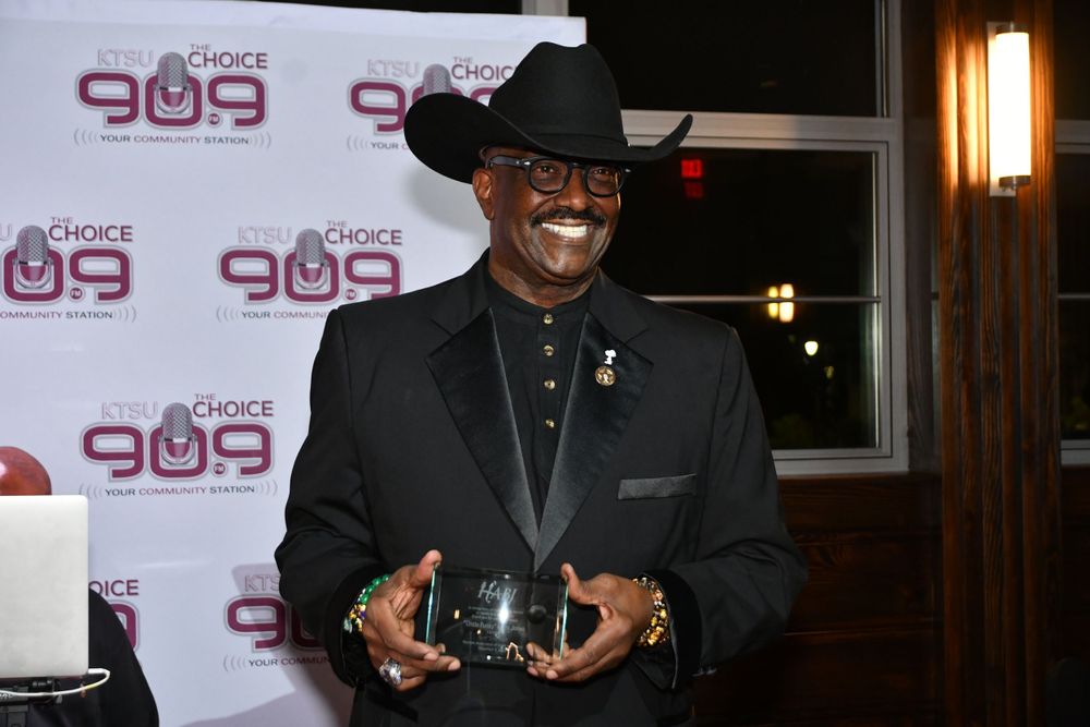 Award recipient in black suit and cowboy hat holding trophy against branded backdrop.