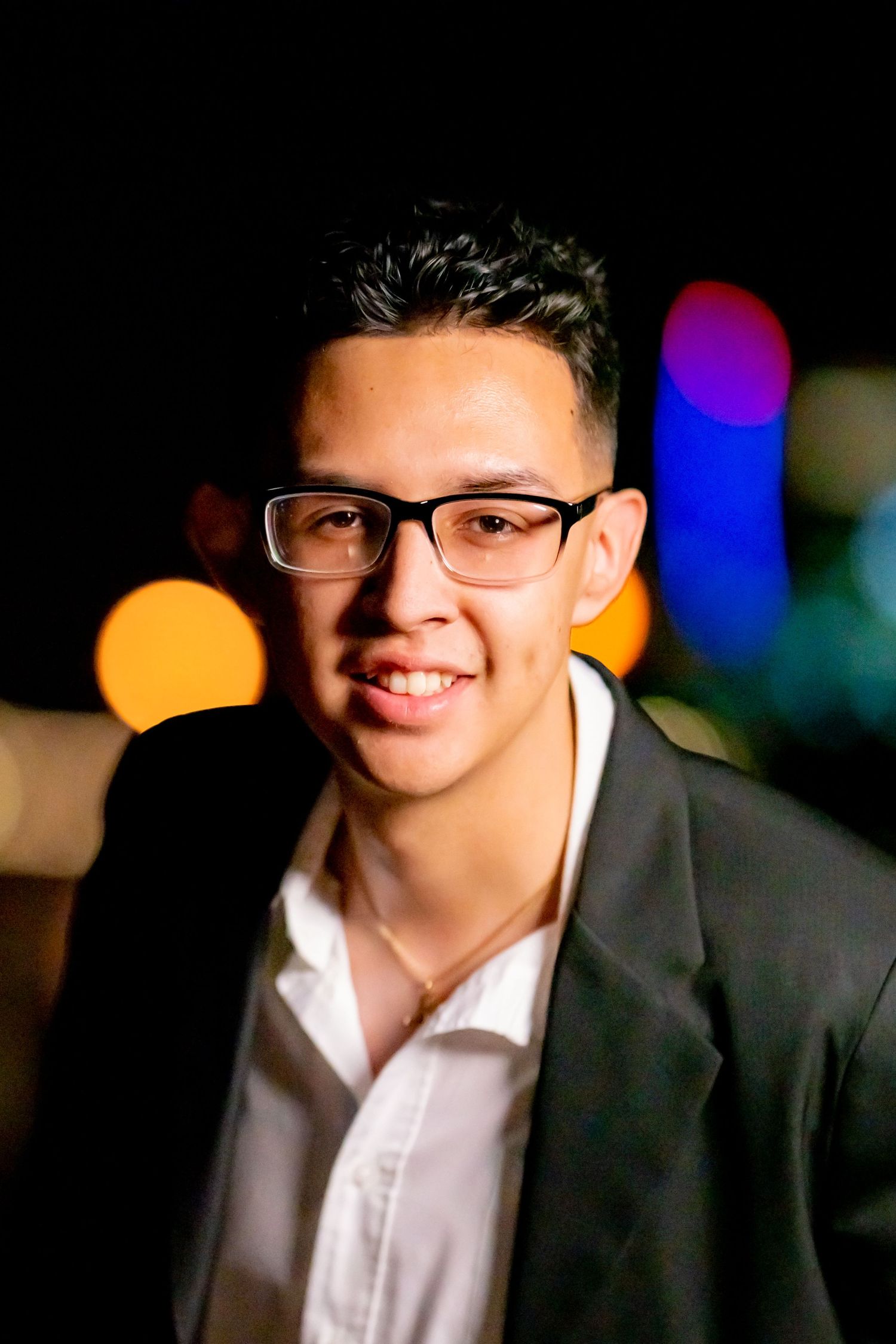 Professional portrait with bokeh lights showing person in glasses and black blazer smiling warmly against dark background.