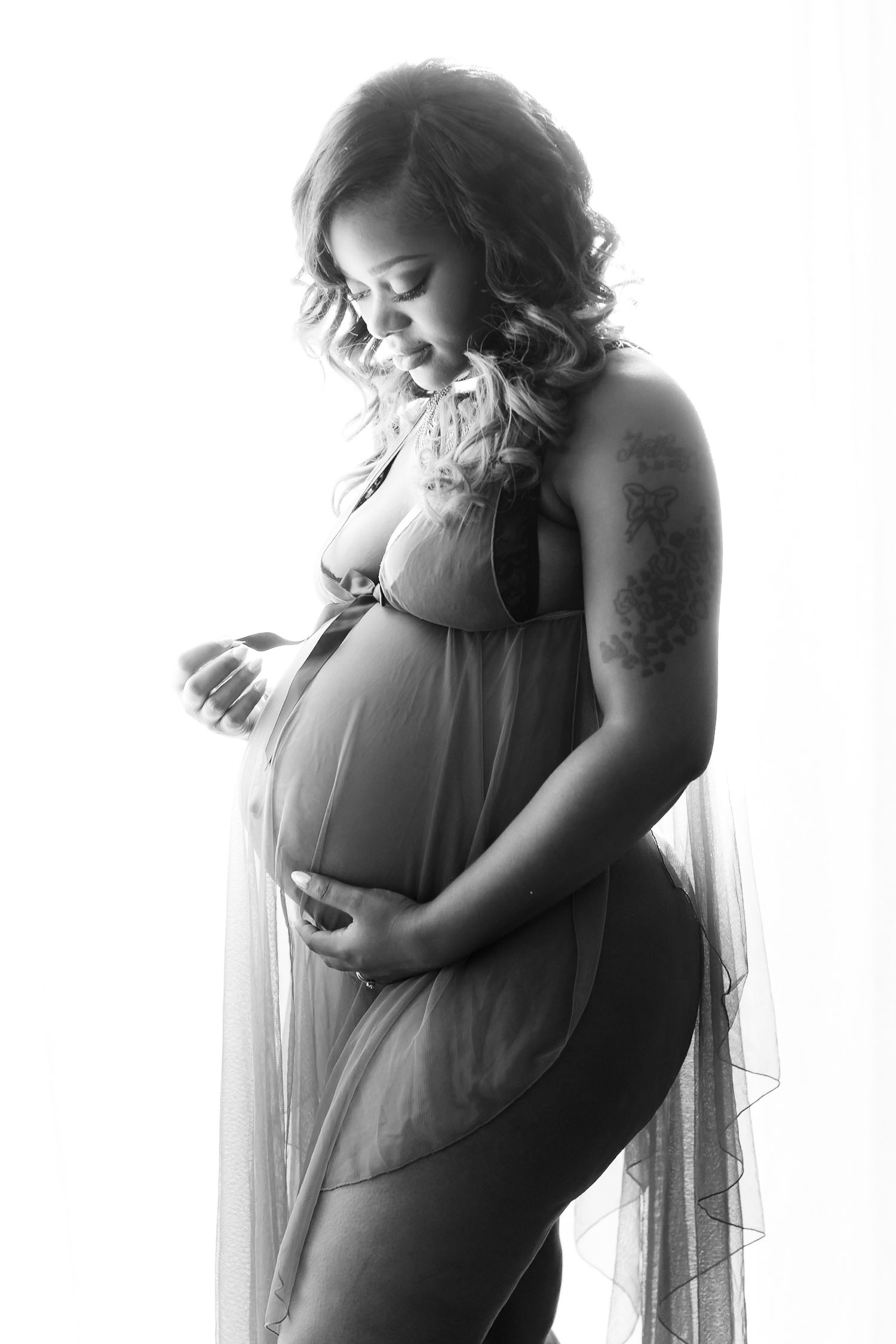 Artistic black and white maternity photograph showing an expectant mother in sheer fabric standing by window light.