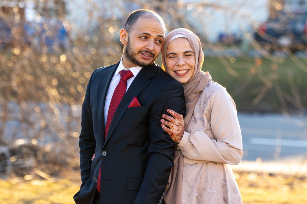 A couple stands close together outdoors, with the person on the right wearing a beige hijab and the person on the left in a suit.
