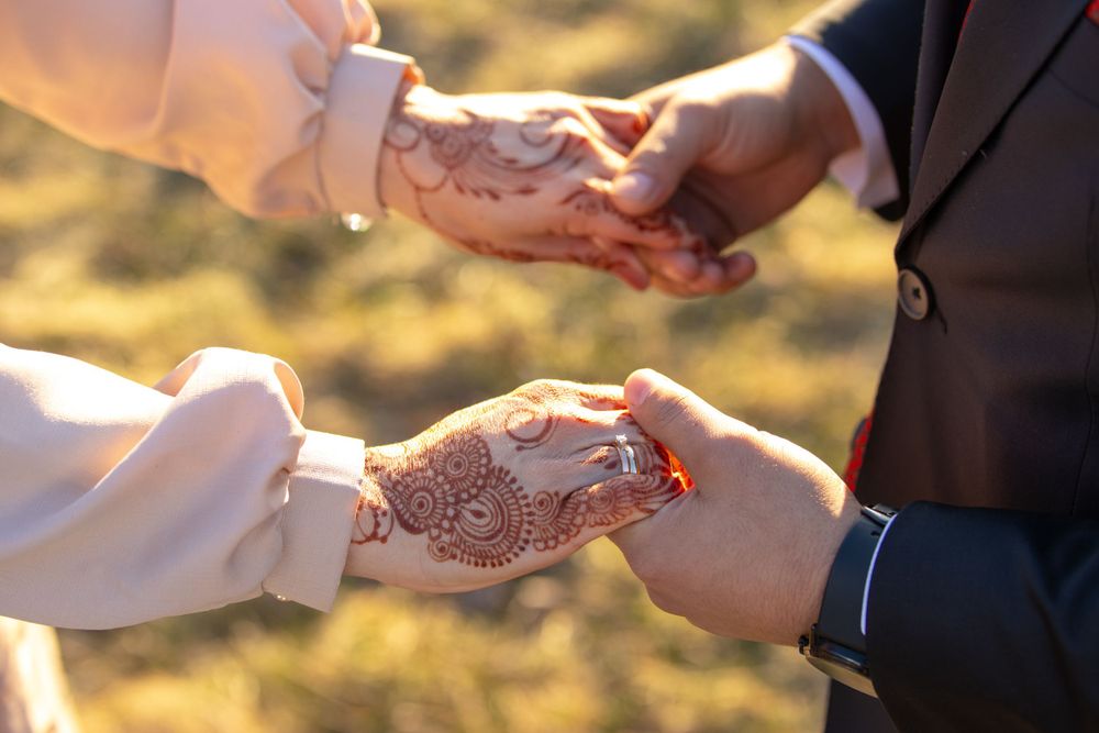 A close up of hands with intricate henna designs exchanging rings during a wedding ceremony at sunset.