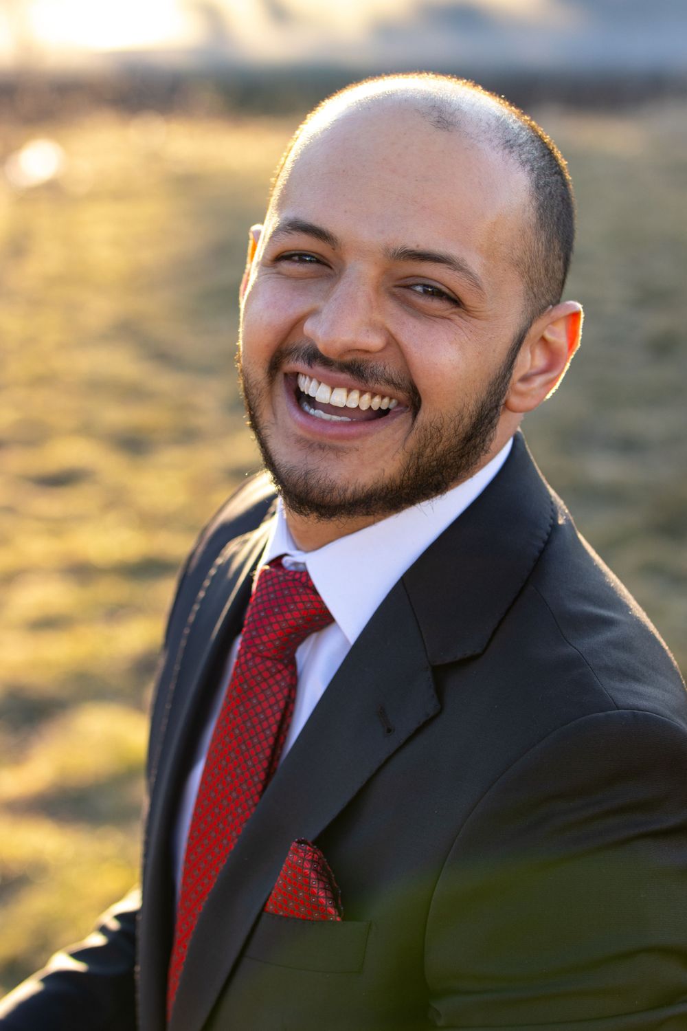 Professional portrait of someone in a black suit with red tie and pocket square smiling warmly in golden outdoor lighting.