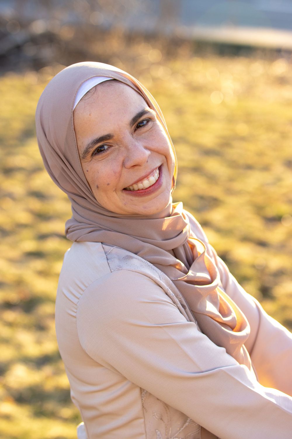A woman wearing a light beige hijab smiles brightly while sitting outdoors in golden sunlight.