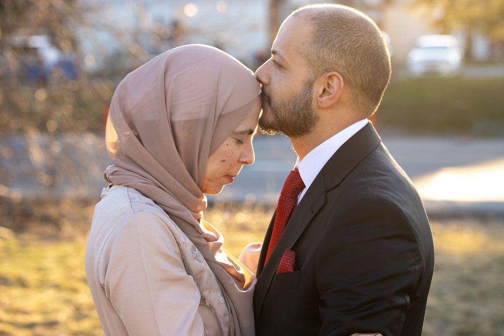 A tender moment between a couple outdoors, with the woman wearing a mauve hijab and the man in a dark suit.