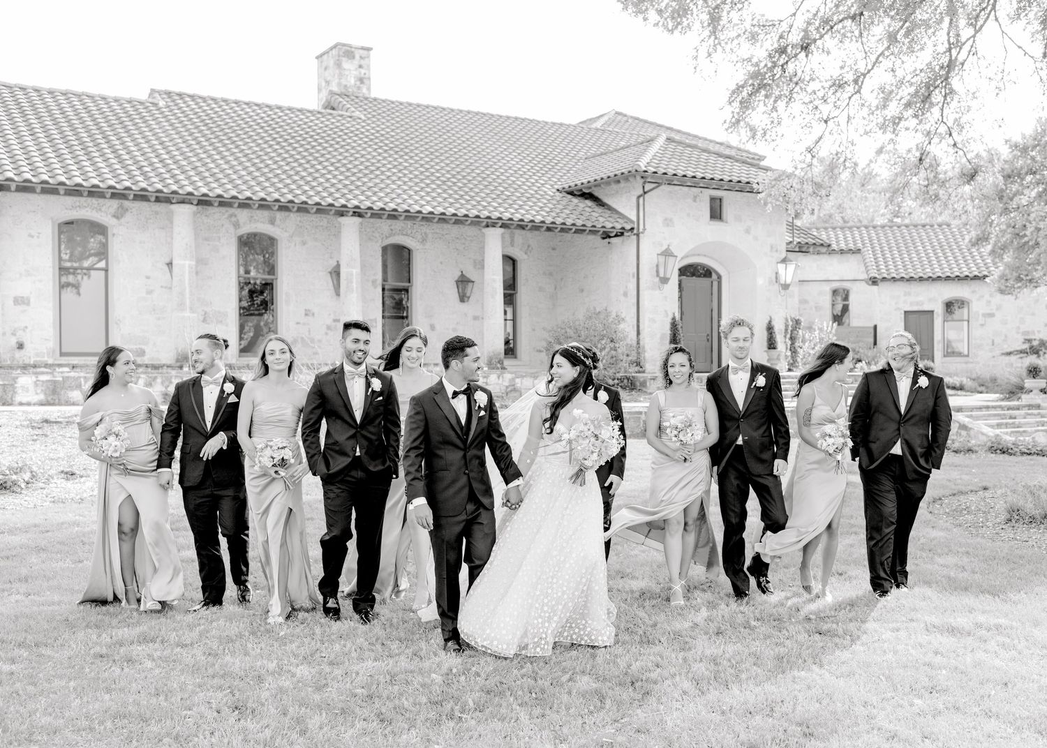 A black and white photo of a wedding party walking and laughing together outside a Spanish-style building.