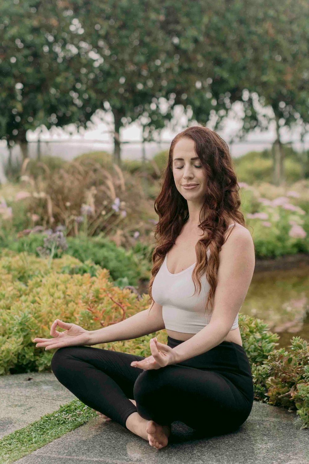 Young woman with auburn hair meditating in lotus pose in a lush garden setting.