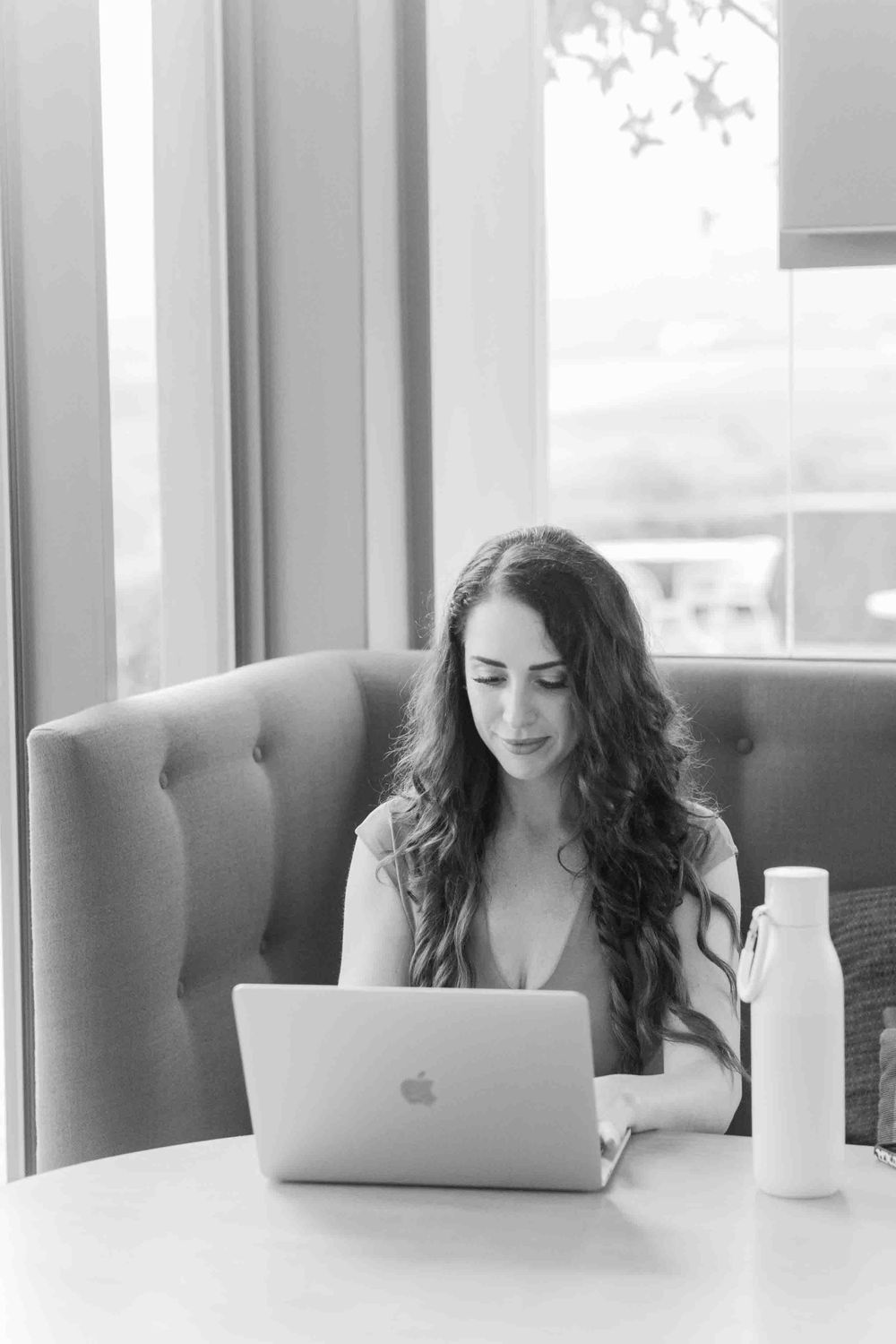 Woman with long wavy hair working on MacBook laptop at white table in bright cafe.