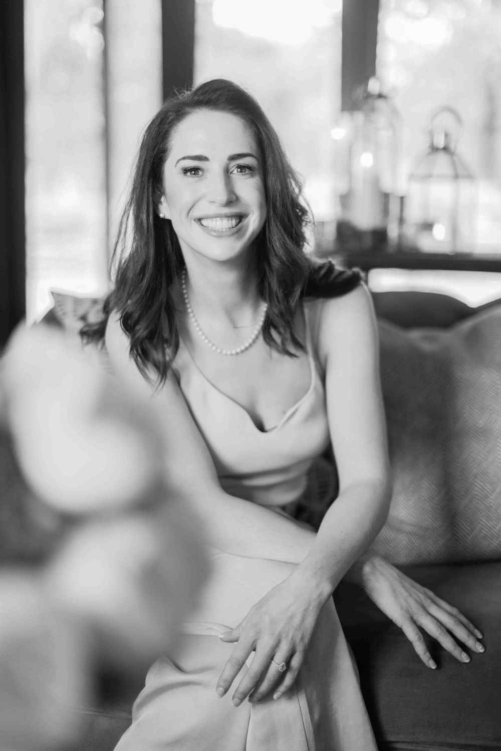 Smiling woman in pearl necklace and slip dress sitting on sofa in black and white portrait.