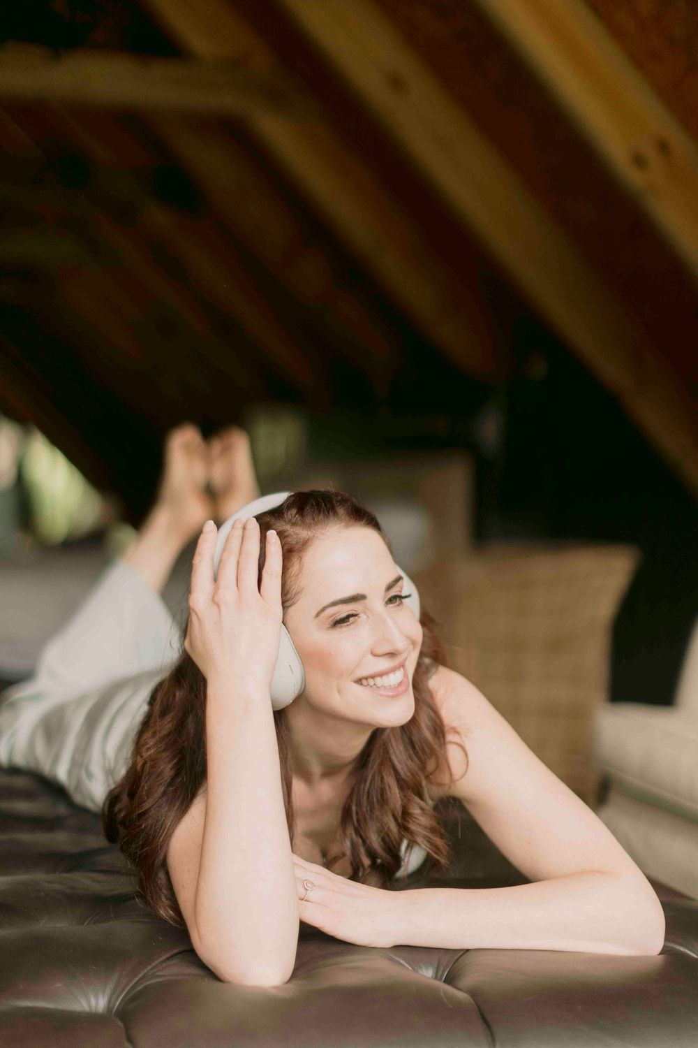 Smiling woman lying on leather sofa wearing white headphones in rustic attic space.