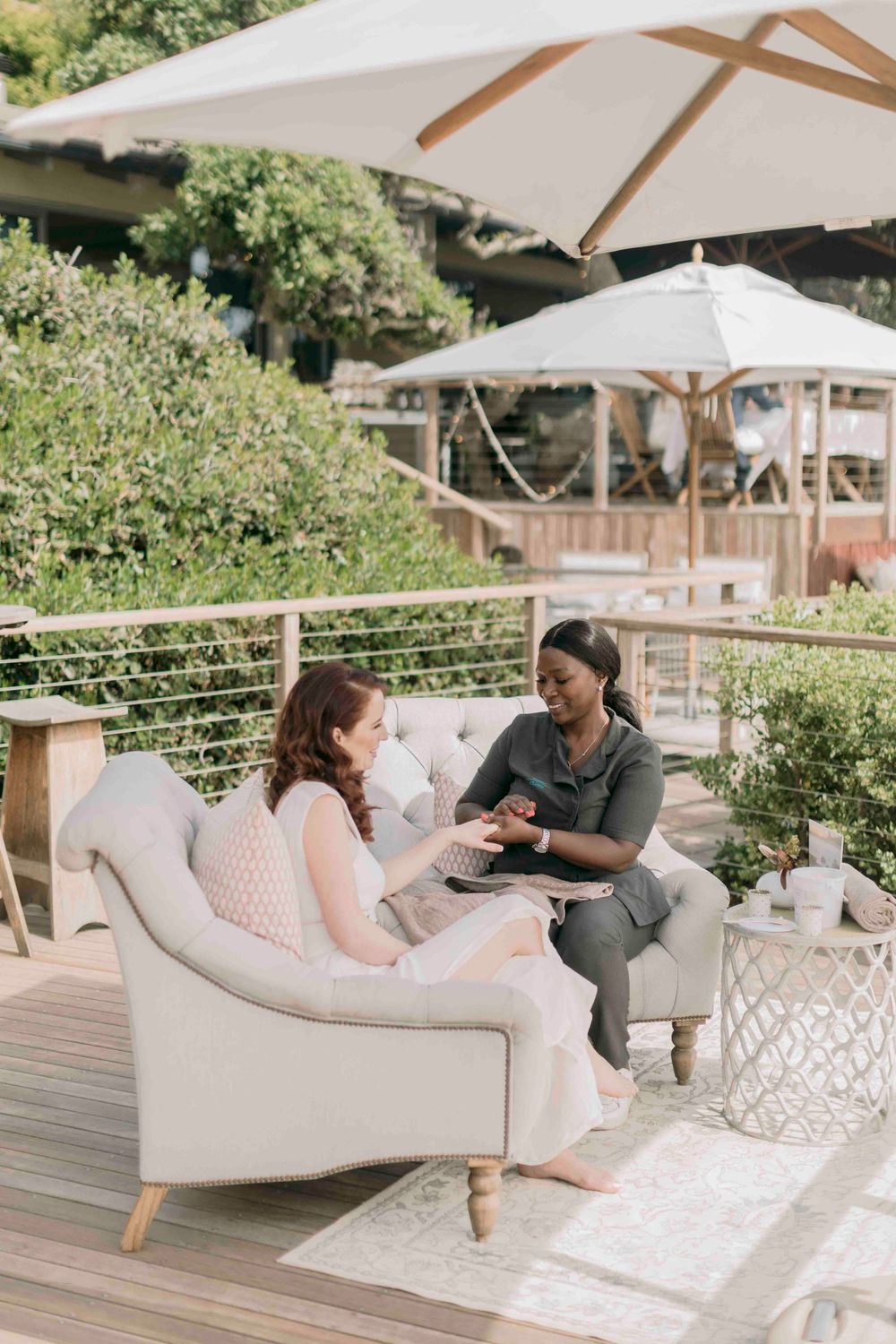 Two women having a relaxed outdoor conversation on elegant patio furniture under a white umbrella.