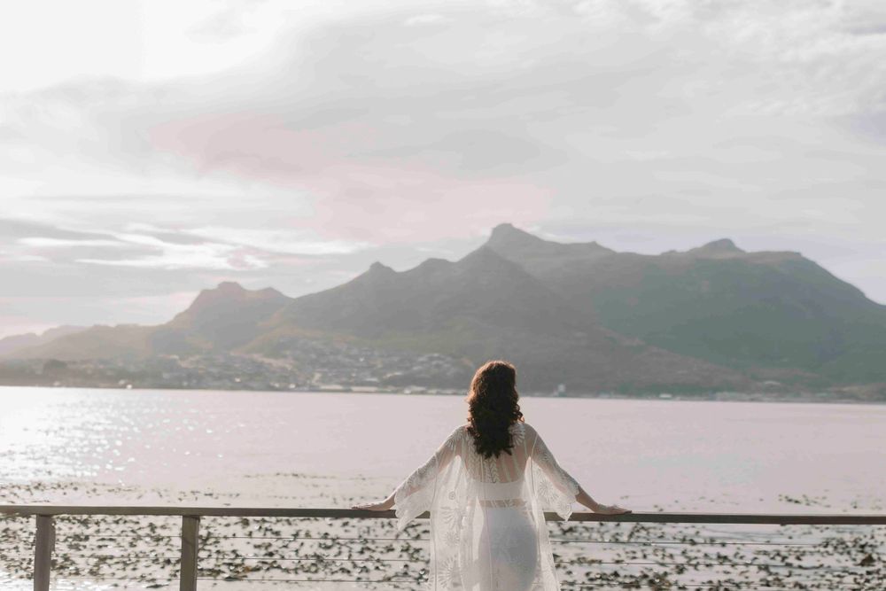 Woman in white lace dress stands at a railing overlooking a calm bay with mountain views.