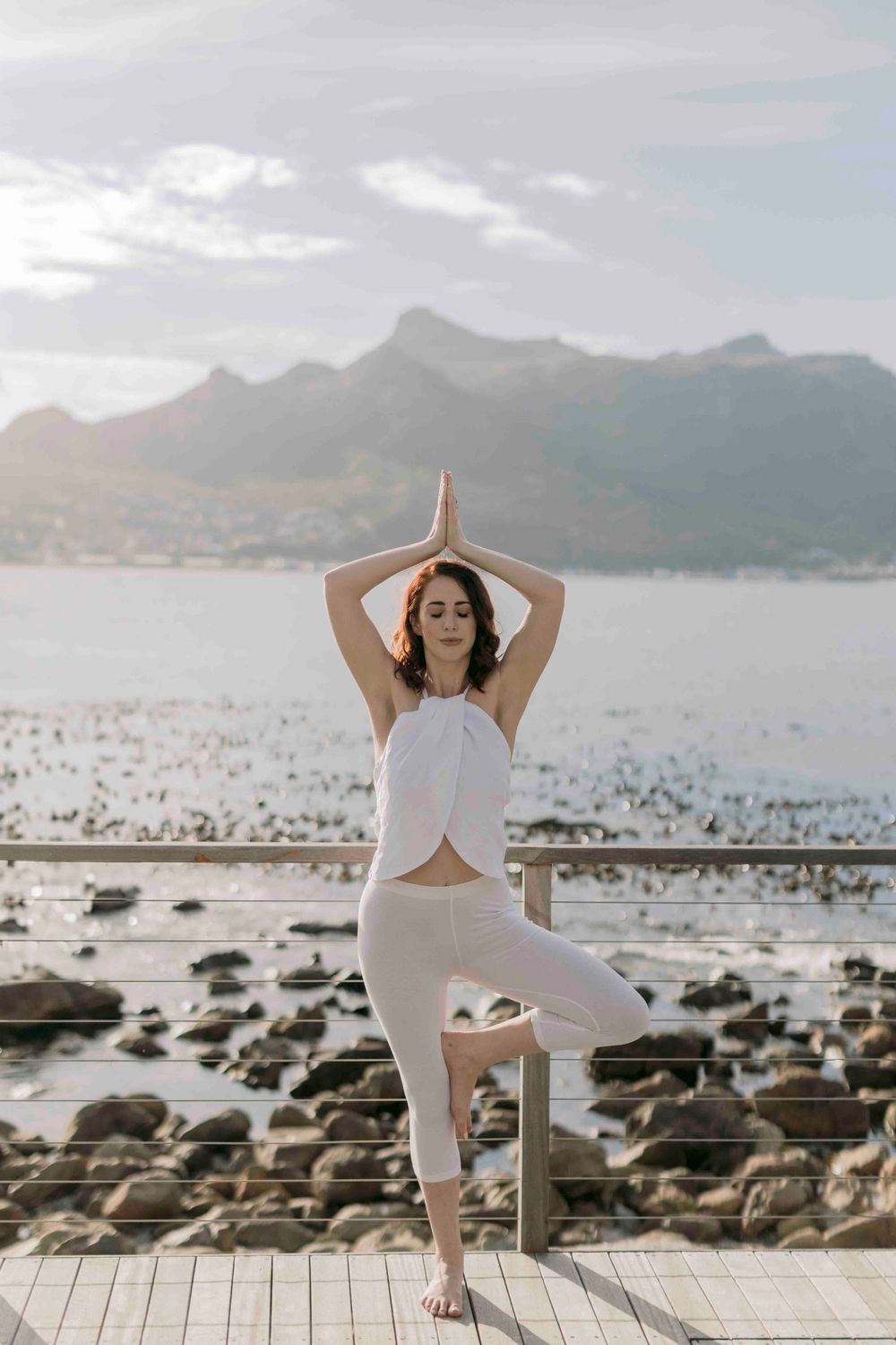 Woman in white performs tree yoga pose on a waterfront deck with mountain scenery at sunrise.