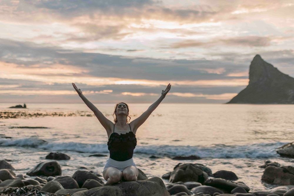 Woman kneeling on rocky beach with arms raised toward dramatic sunset sky and ocean mountain view.