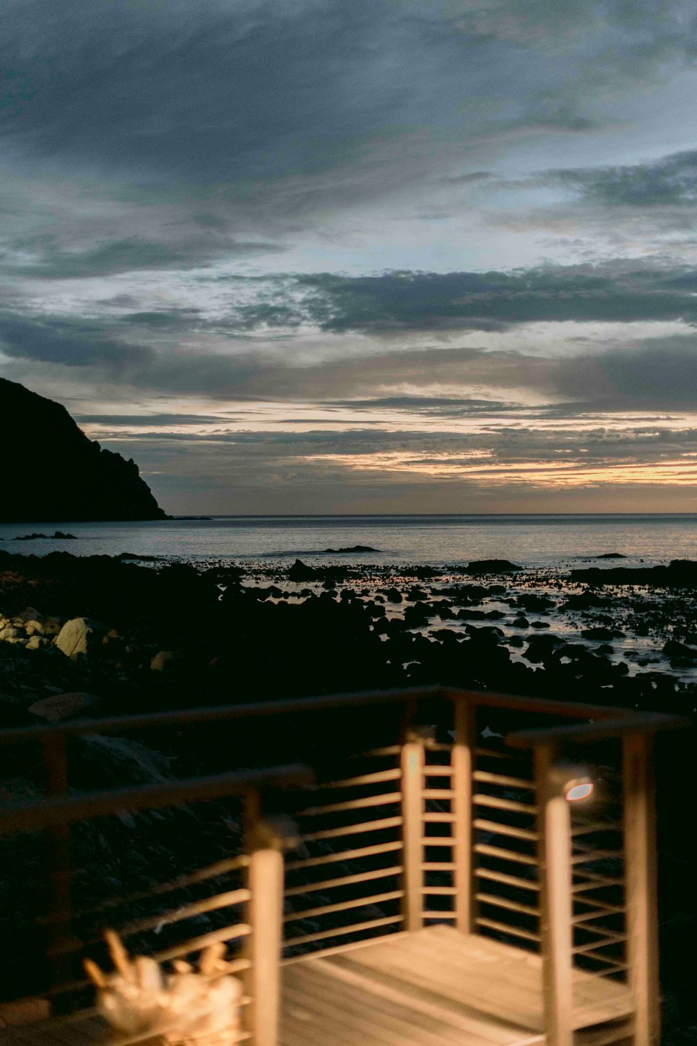 Wooden deck overlooking rocky coastline at dusk with glowing sunset horizon and dramatic cloudy sky.