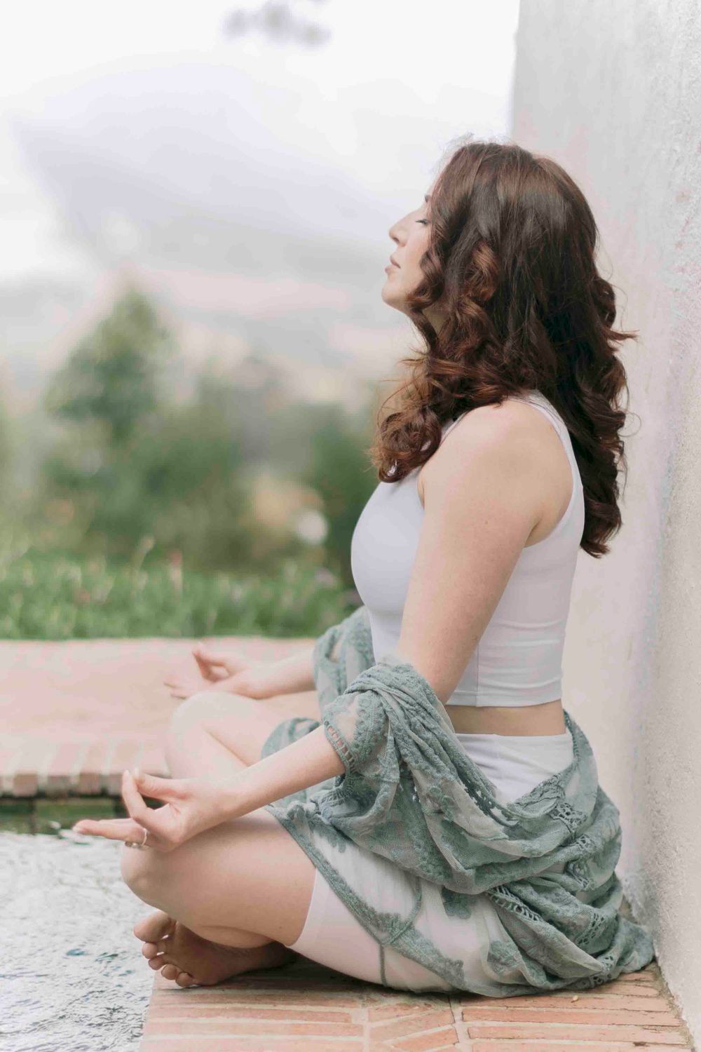 Woman sitting cross-legged in meditation with eyes closed against a white wall in a serene outdoor setting.