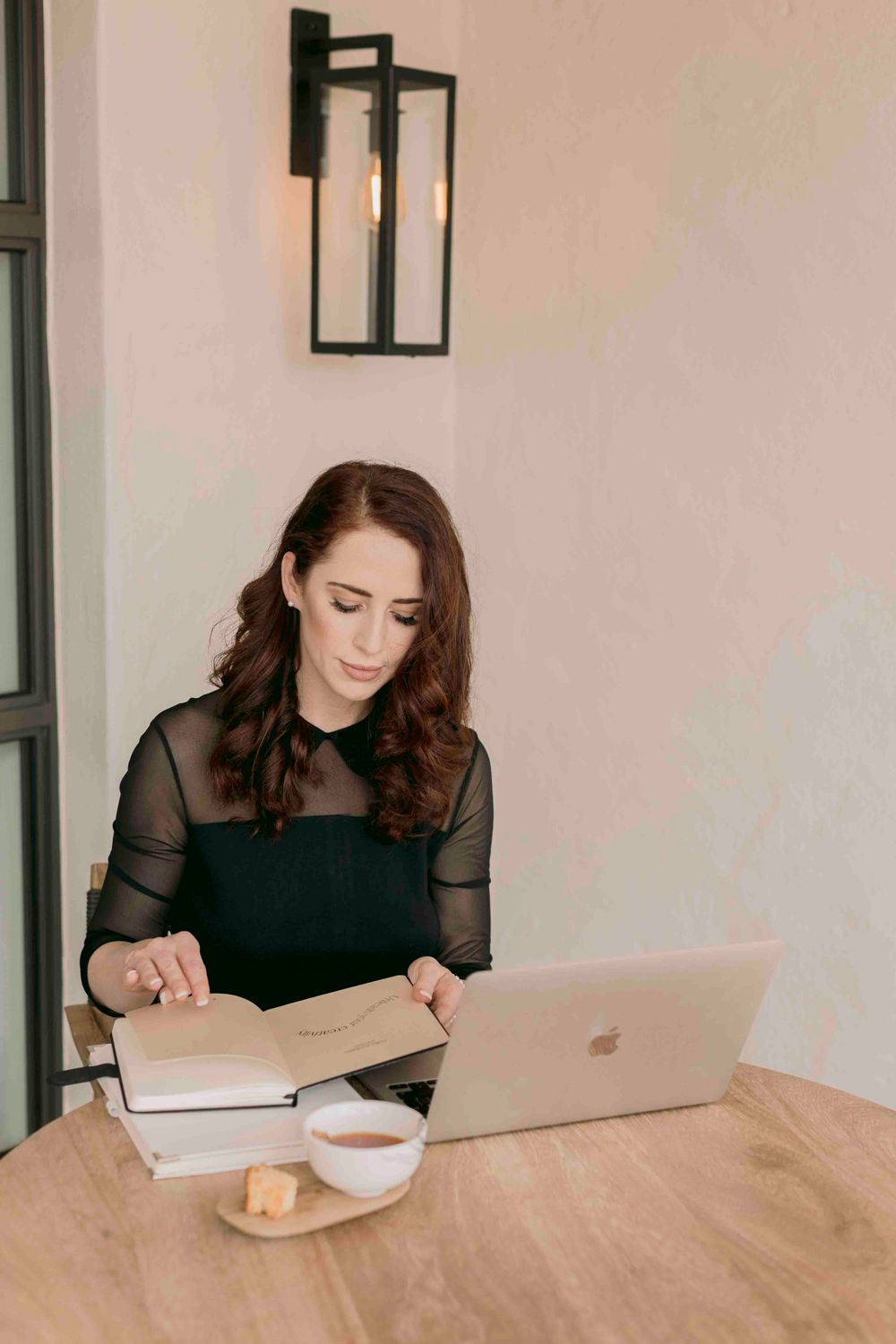 Brunette woman in black mesh top working at wooden desk with MacBook and notebook.