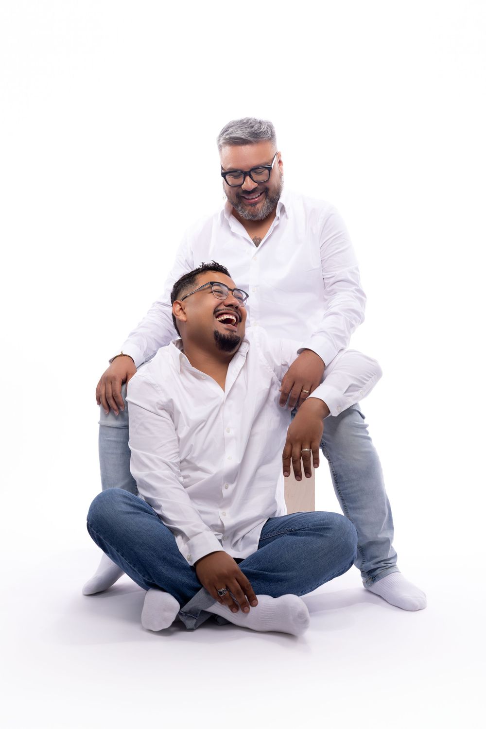 Two people wearing white shirts and jeans sit and stand together in a playful pose against white background.