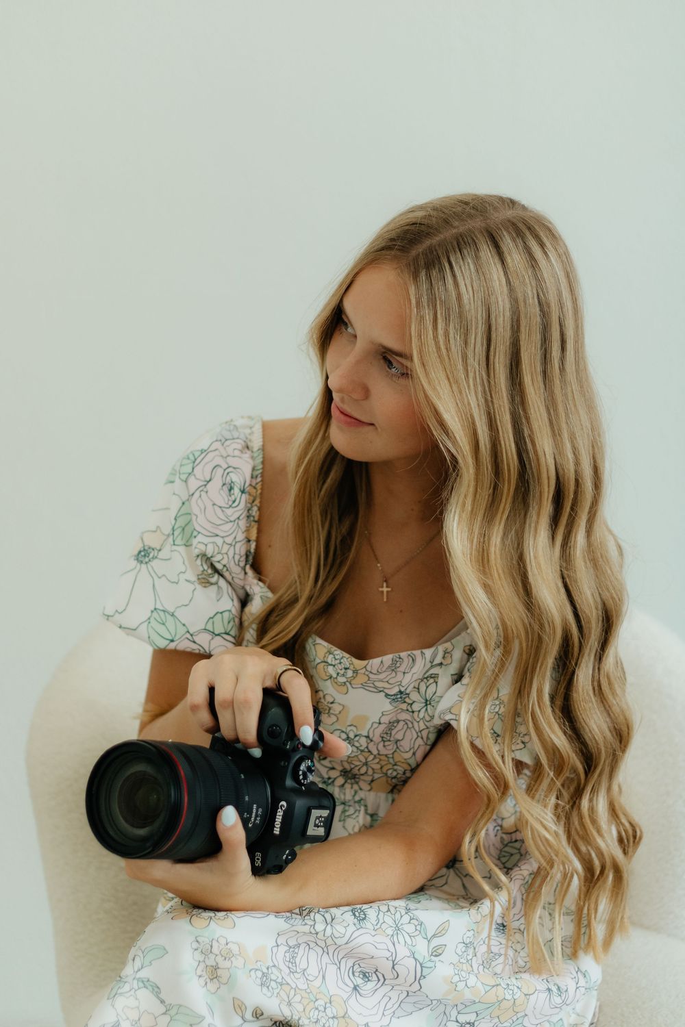 Person with wavy blonde hair wearing a floral dress holds a black camera while posing against a light background.