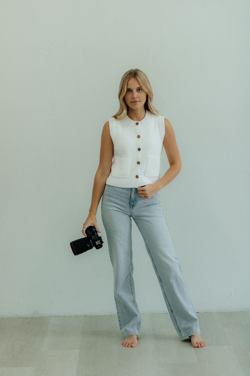 A woman in a white sleeveless top and light blue wide-leg pants poses against a neutral wall with a camera.