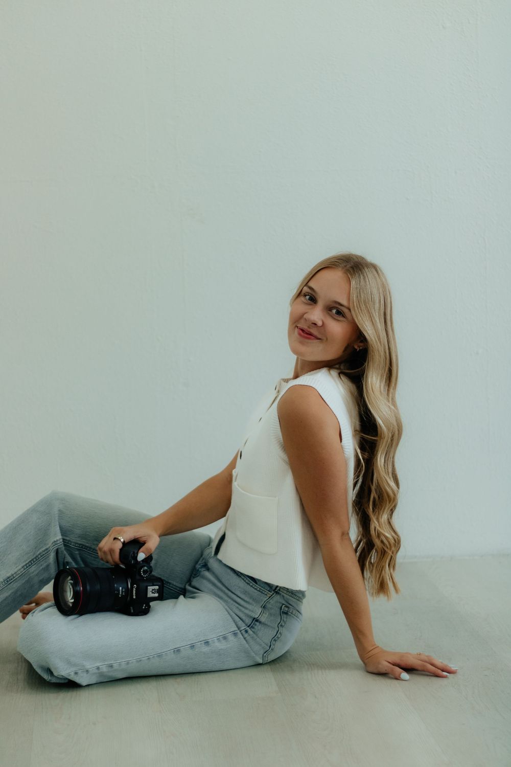 A person in a white tank top and light jeans poses on the floor against a white background in a minimalist photoshoot.