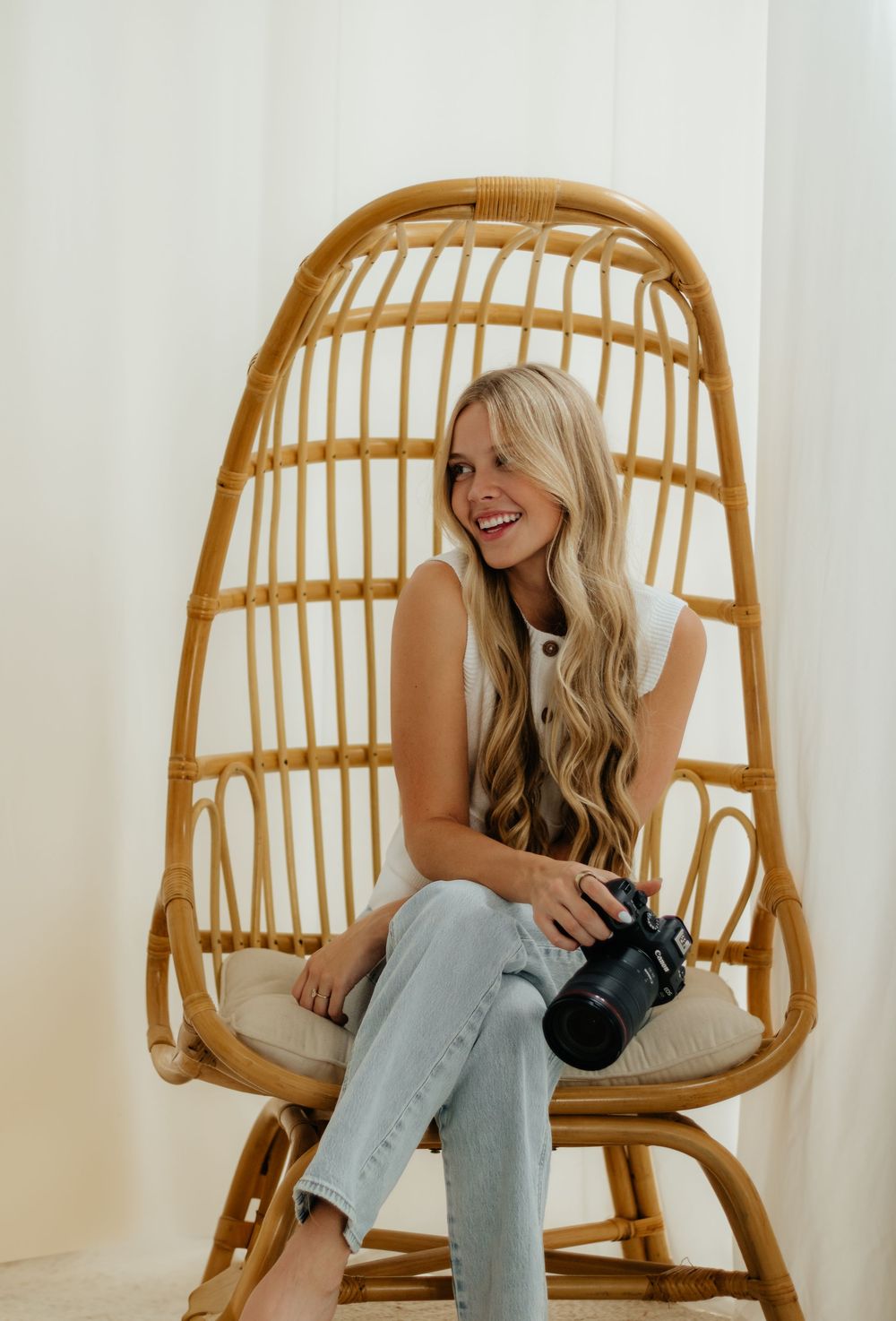 A person sits and laughs joyfully in a rattan hanging chair against a white wall.