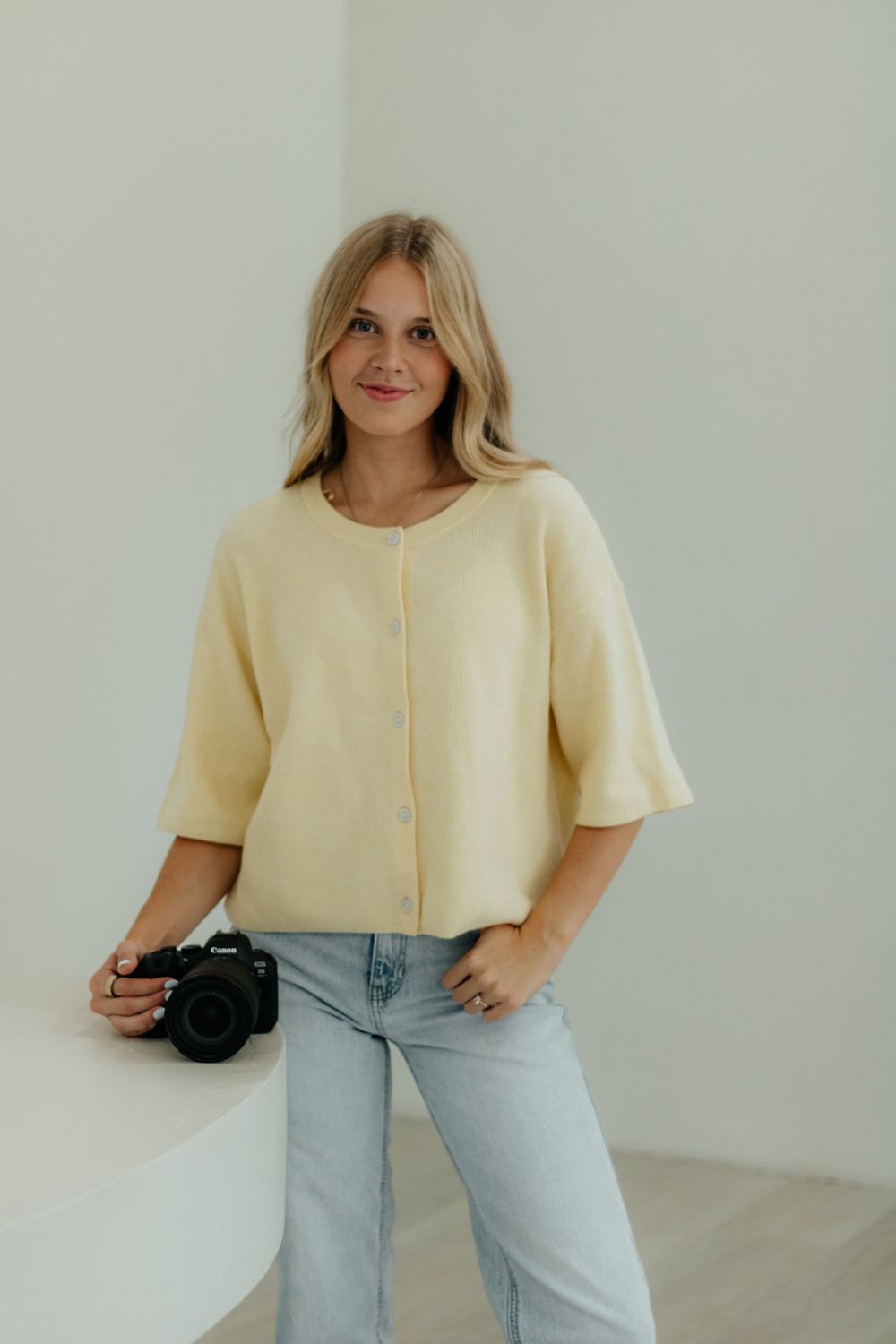 Woman in cream colored crop top and light wash jeans poses against white wall in minimalist fashion photoshoot.