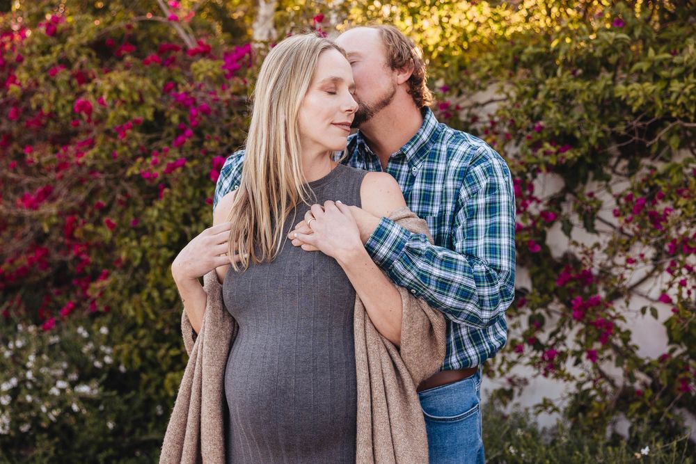 A tender maternity moment captured outdoors with blooming flowers and greenery in the background.