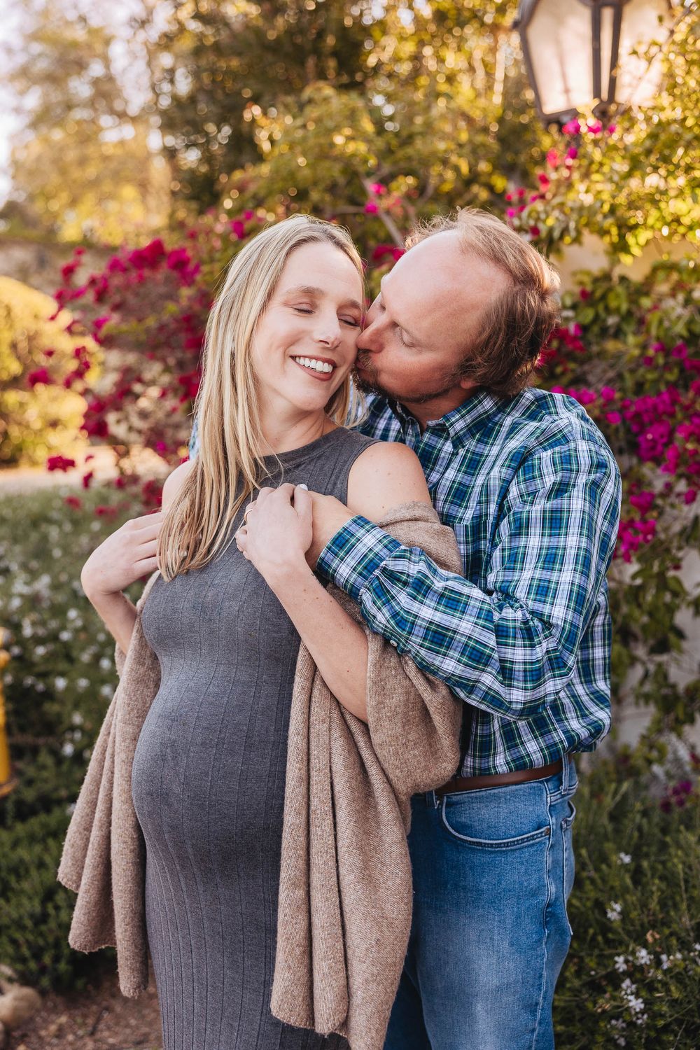 A romantic embrace in a garden with blooming pink flowers and warm sunlit trees in the background.