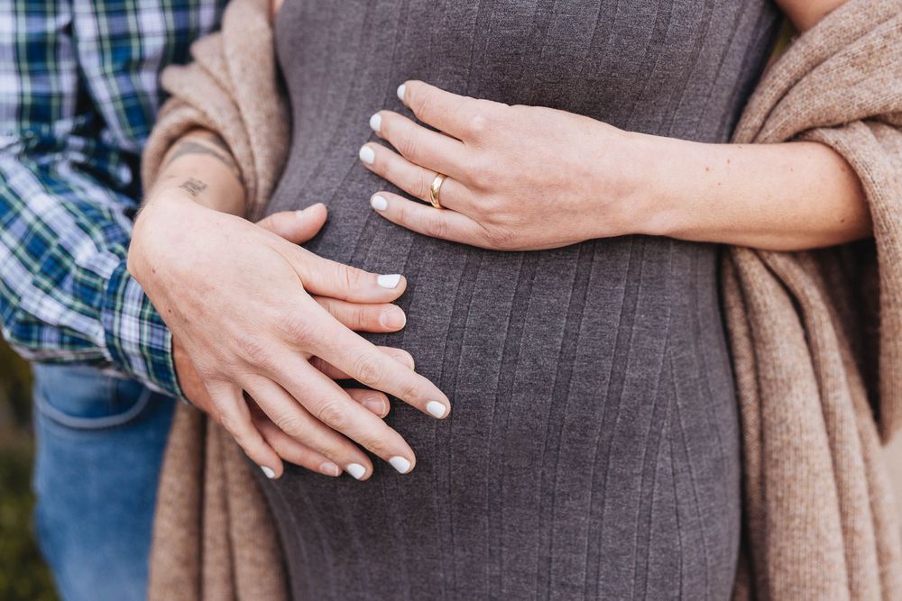 Close up of hands resting on a pregnant belly, wearing a brown sweater and tan cardigan.