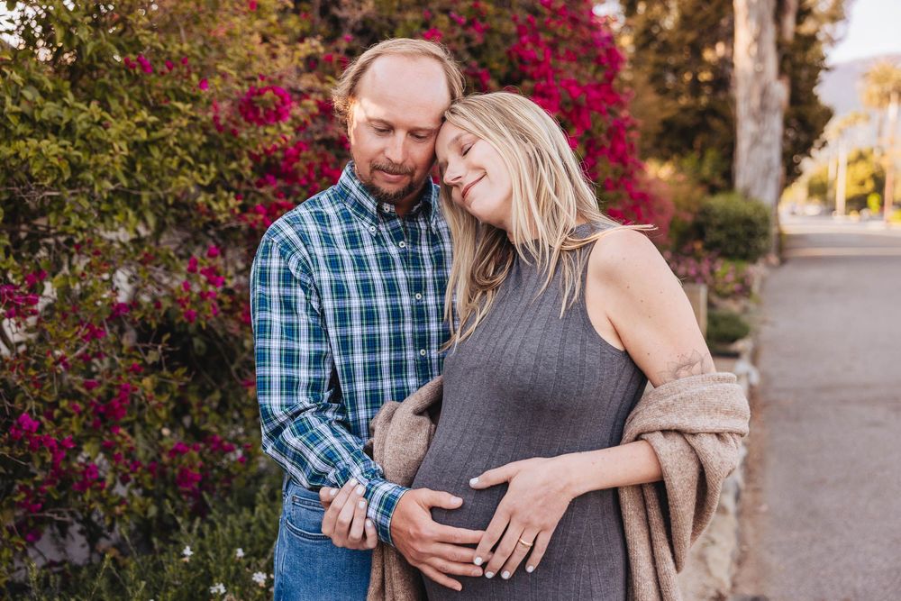 Couple poses together in front of blooming red bougainvillea during a maternity photoshoot outdoors.
