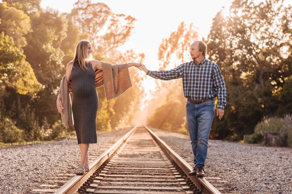 A couple holds hands while balancing on railroad tracks as golden sunlight streams through trees in the background.