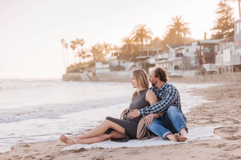 A couple sits close together on a sandy beach at sunset with coastal homes and palm trees in the background.