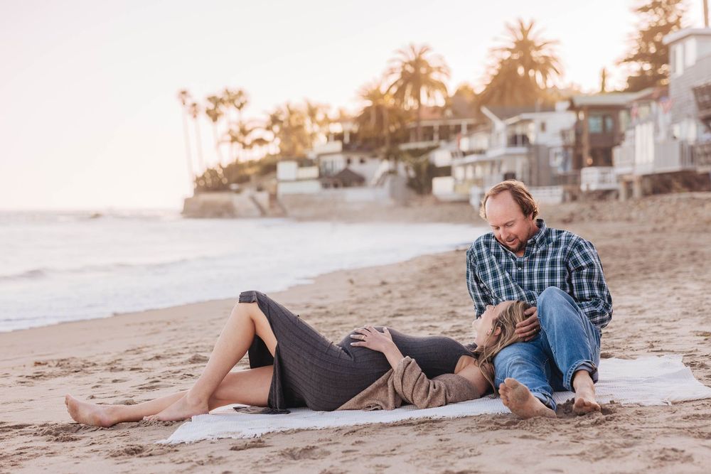 A couple relaxes on a blanket at sunset on a beach with coastal homes visible in the background.