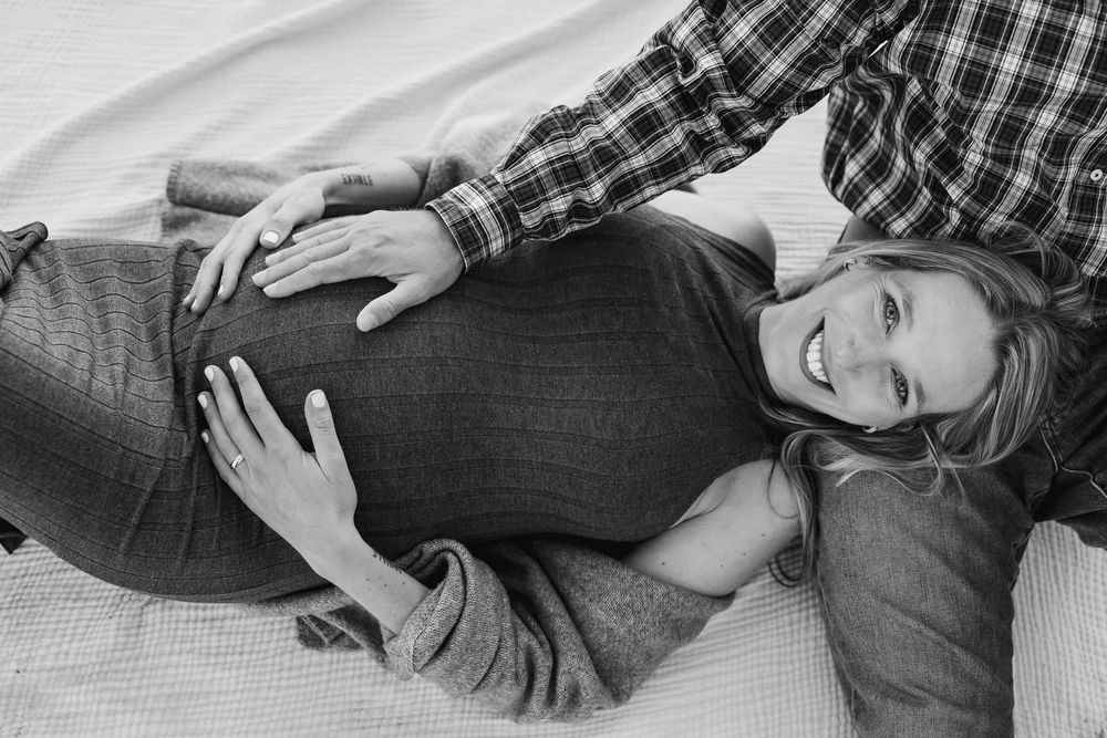A person wearing a plaid shirt rests on white bedding while cradling their pregnant belly in a candid black and white photo.