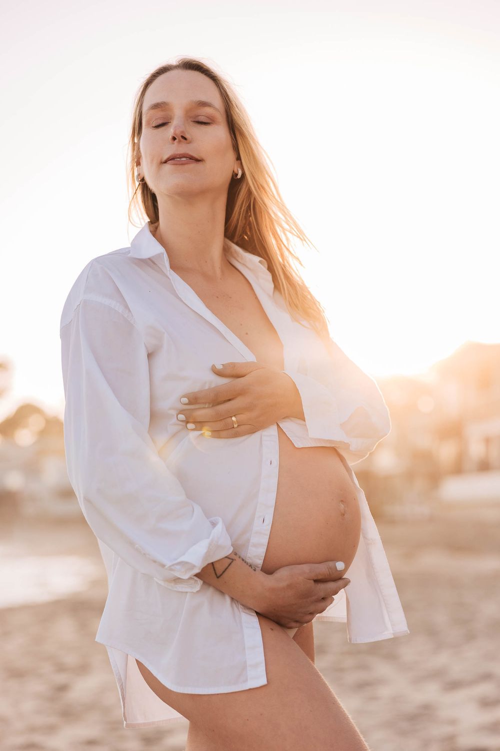 Expectant mother in white button down shirt poses on beach during golden hour.