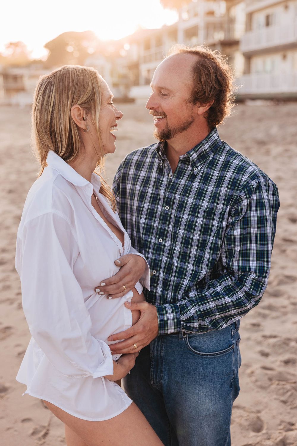 Joyful couple shares intimate moment on beach during maternity photoshoot at sunset.